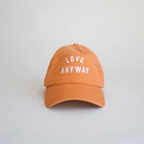 An orange baseball cap with the text 'LOVE ANYWAY' printed in white on the front.