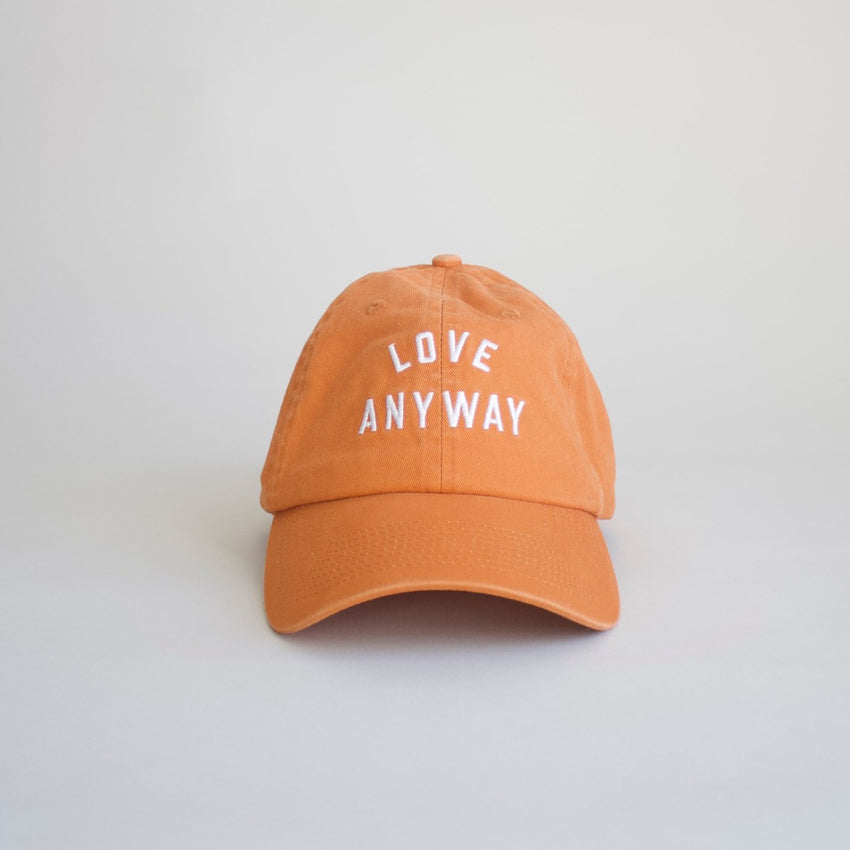 An orange baseball cap with the text 'LOVE ANYWAY' printed in white on the front.